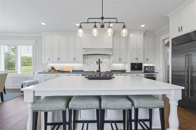 kitchen with a breakfast bar, built in fridge, a center island with sink, decorative light fixtures, and white cabinetry