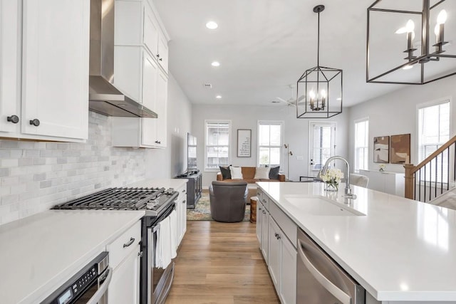 kitchen with sink, decorative light fixtures, a center island with sink, appliances with stainless steel finishes, and wall chimney range hood