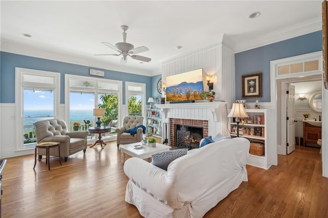 living area featuring a wainscoted wall, a fireplace, wood finished floors, a ceiling fan, and ornamental molding