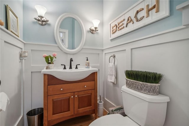 half bath with toilet, wainscoting, a decorative wall, and vanity