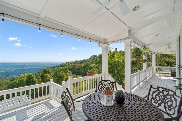 wooden deck with a forest view, ceiling fan, a porch, and outdoor dining space