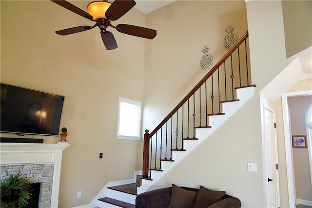 staircase with a stone fireplace, a ceiling fan, and a towering ceiling