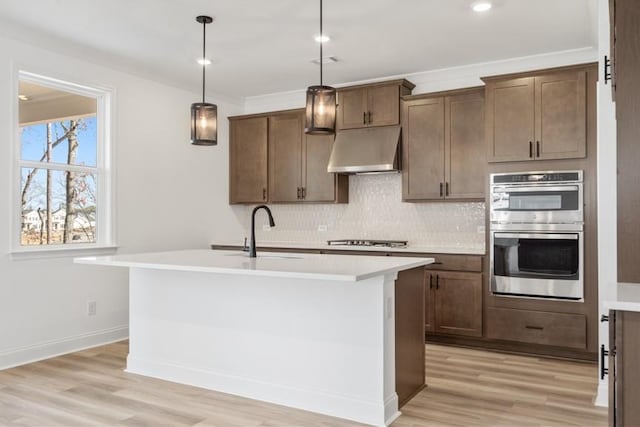 kitchen with stainless steel appliances, light countertops, hanging light fixtures, an island with sink, and under cabinet range hood