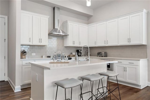 kitchen featuring wall chimney range hood, decorative backsplash, a kitchen island with sink, a sink, and dark wood-style floors