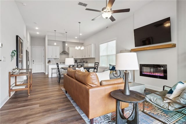 living area featuring dark wood-style flooring, visible vents, baseboards, ceiling fan with notable chandelier, and a glass covered fireplace