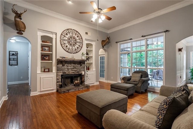 living room featuring a stone fireplace, crown molding, built in shelves, ceiling fan, and wood-type flooring