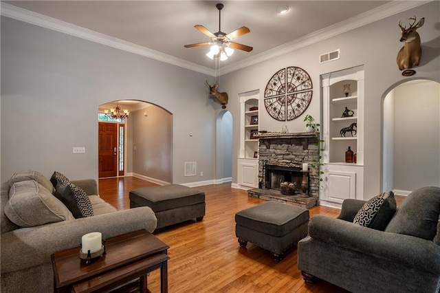 living room featuring built in shelves, a stone fireplace, hardwood / wood-style floors, ceiling fan with notable chandelier, and ornamental molding