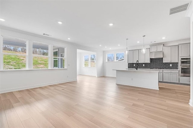 kitchen with hanging light fixtures, light hardwood / wood-style flooring, double oven, a kitchen island with sink, and decorative backsplash