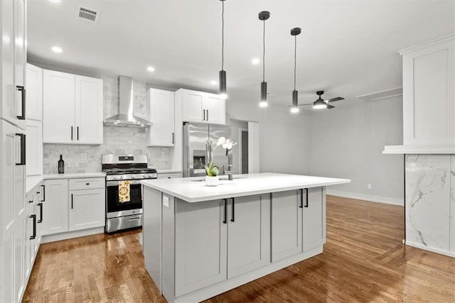 kitchen featuring ceiling fan, wall chimney exhaust hood, an island with sink, white cabinets, and appliances with stainless steel finishes