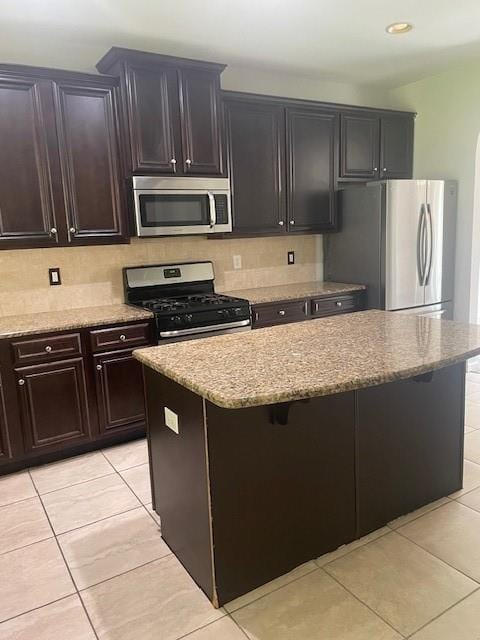 kitchen featuring dark brown cabinetry, light tile patterned floors, stainless steel appliances, light stone countertops, and backsplash