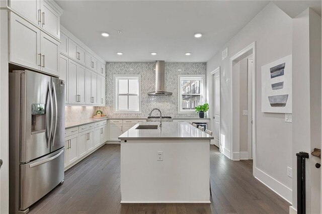 kitchen with stainless steel fridge, wall chimney exhaust hood, dark hardwood / wood-style floors, white cabinetry, and an island with sink