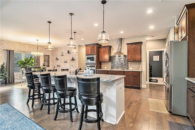 kitchen with wall chimney exhaust hood, appliances with stainless steel finishes, a kitchen island with sink, and pendant lighting