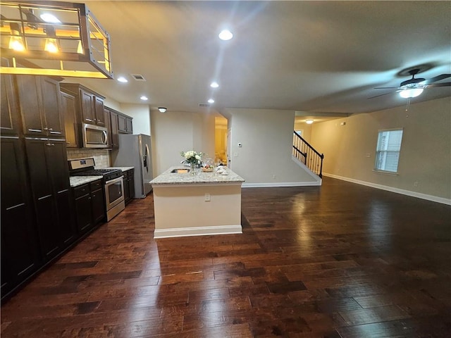 kitchen featuring dark wood-type flooring, stainless steel appliances, light stone countertops, dark brown cabinets, and a center island with sink