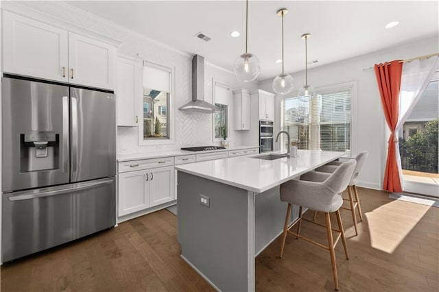 kitchen with white cabinetry, decorative light fixtures, stainless steel appliances, a kitchen island with sink, and wall chimney range hood