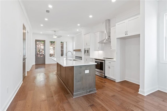 kitchen with wall chimney range hood, white cabinets, an island with sink, and stainless steel appliances