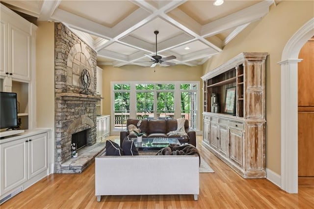 living room featuring coffered ceiling, a stone fireplace, ceiling fan, light wood-type flooring, and beam ceiling