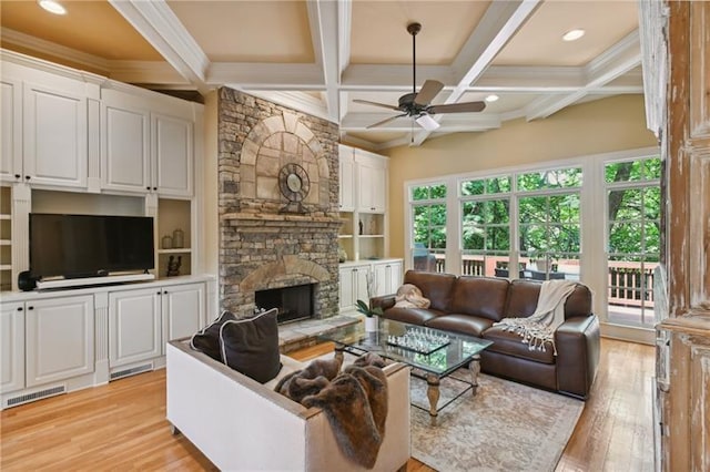 living room featuring ceiling fan, beam ceiling, coffered ceiling, light hardwood / wood-style flooring, and a stone fireplace