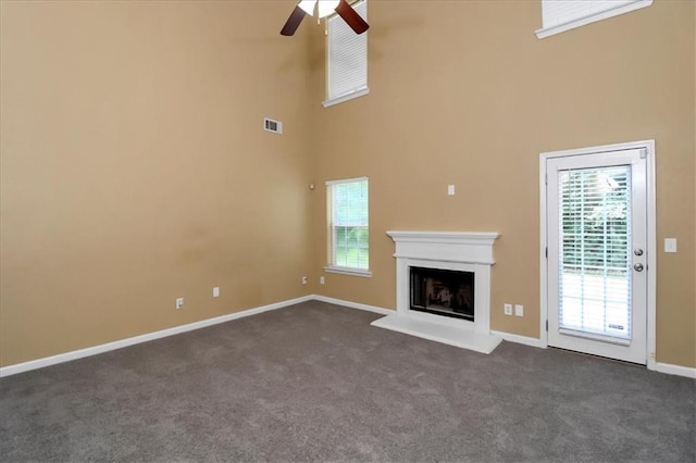 unfurnished living room featuring visible vents, a fireplace with raised hearth, dark carpet, a high ceiling, and a ceiling fan