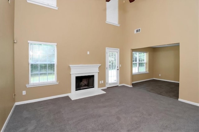 unfurnished living room with visible vents, a fireplace with raised hearth, dark carpet, a high ceiling, and ceiling fan