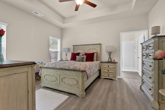 bedroom with a raised ceiling, light wood-style flooring, a ceiling fan, baseboards, and connected bathroom