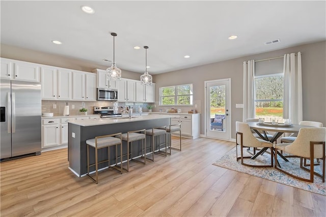 kitchen with appliances with stainless steel finishes, hanging light fixtures, white cabinetry, light hardwood / wood-style floors, and a center island with sink