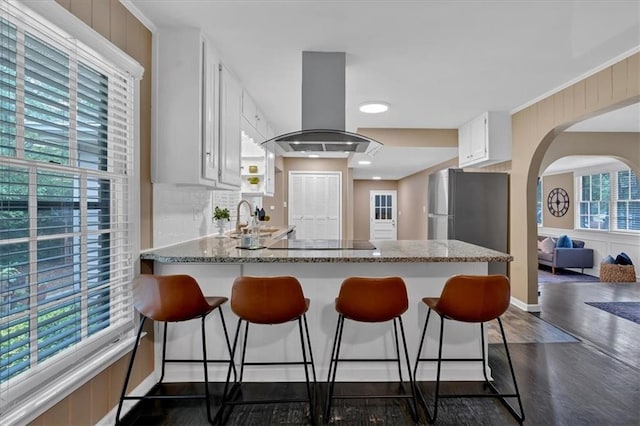 kitchen featuring stainless steel refrigerator, light stone countertops, island exhaust hood, black electric stovetop, and white cabinets