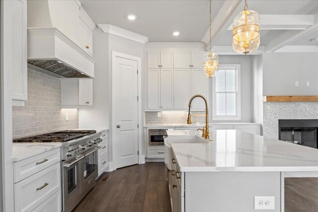 kitchen featuring beam ceiling, range with two ovens, white cabinetry, hanging light fixtures, and an island with sink