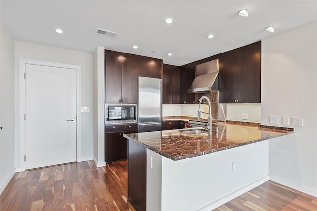 kitchen with built in appliances, kitchen peninsula, hardwood / wood-style floors, and wall chimney range hood