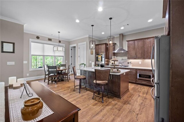 kitchen with wall chimney range hood, appliances with stainless steel finishes, pendant lighting, light hardwood / wood-style flooring, and a kitchen island with sink