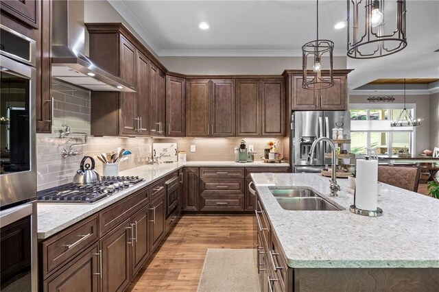 kitchen with wall chimney range hood, an island with sink, sink, crown molding, and decorative light fixtures