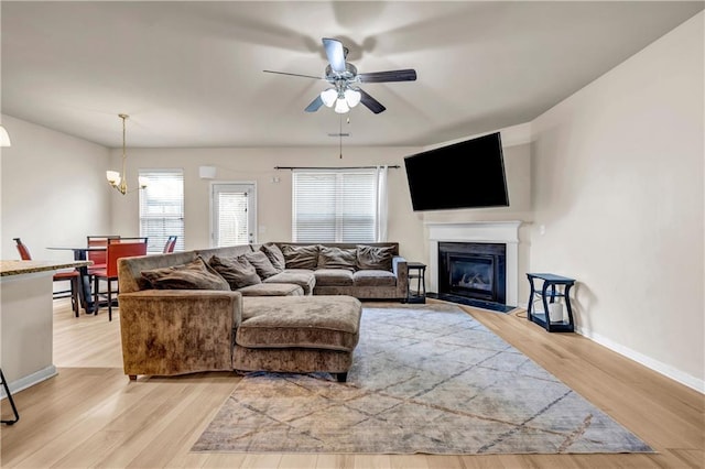 living area with ceiling fan with notable chandelier, a glass covered fireplace, light wood-style flooring, and baseboards