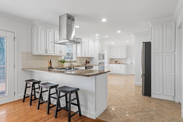 kitchen featuring appliances with stainless steel finishes, island range hood, white cabinetry, dark stone countertops, and kitchen peninsula