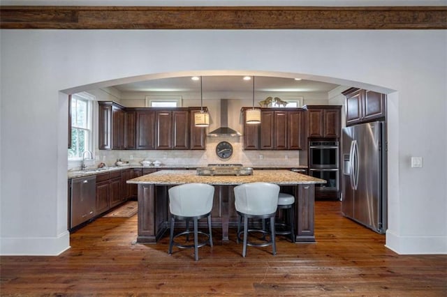 kitchen featuring wall chimney exhaust hood, stainless steel appliances, a center island, and hanging light fixtures