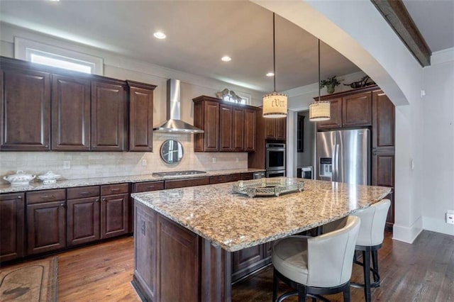 kitchen featuring pendant lighting, a center island, stainless steel appliances, dark brown cabinets, and wall chimney range hood