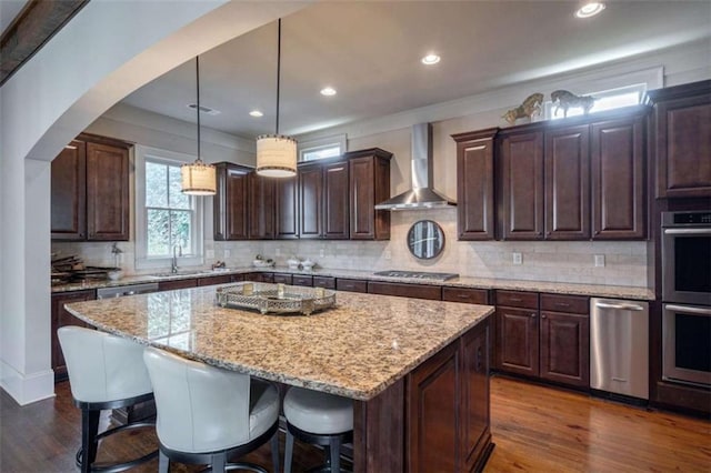 kitchen with decorative light fixtures, a center island, stainless steel appliances, dark brown cabinets, and wall chimney range hood