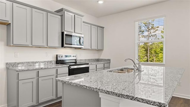 kitchen featuring stainless steel appliances, a sink, gray cabinetry, a kitchen island with sink, and light stone countertops