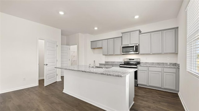 kitchen with appliances with stainless steel finishes, gray cabinetry, a kitchen island with sink, dark wood-style floors, and light stone counters
