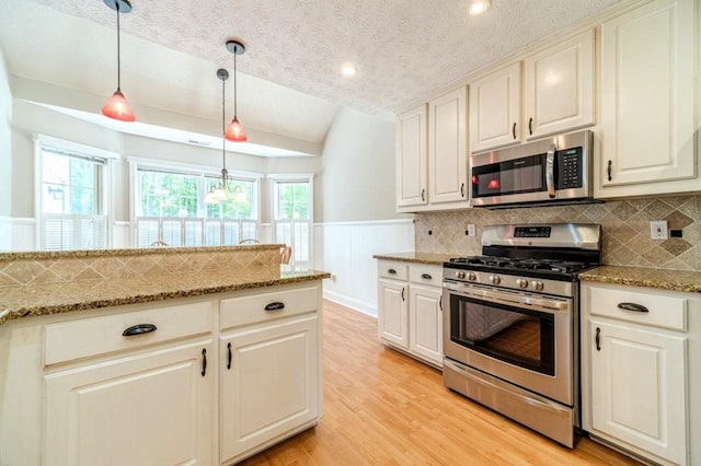 kitchen featuring lofted ceiling, decorative light fixtures, light wood finished floors, a wainscoted wall, and appliances with stainless steel finishes
