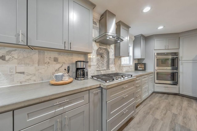 kitchen featuring decorative backsplash, appliances with stainless steel finishes, wall chimney exhaust hood, gray cabinetry, and light hardwood / wood-style flooring