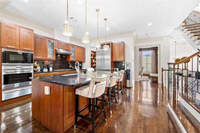 kitchen featuring brown cabinetry, an island with sink, built in appliances, under cabinet range hood, and a warming drawer