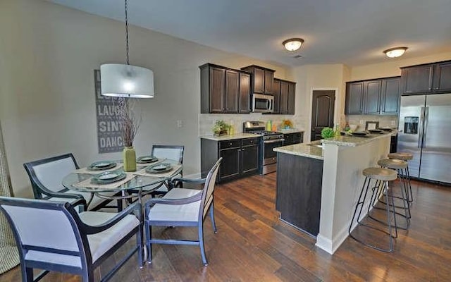 kitchen with pendant lighting, dark wood-type flooring, appliances with stainless steel finishes, dark brown cabinets, and a center island with sink