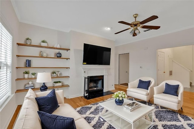 living area with ceiling fan, light wood finished floors, a fireplace, and crown molding