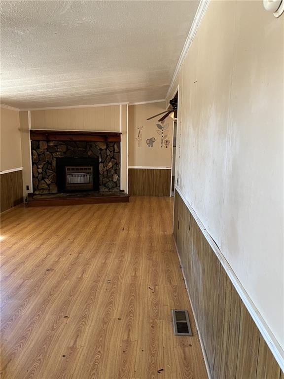 unfurnished living room featuring a stone fireplace, a textured ceiling, light wood-type flooring, ornamental molding, and wooden walls