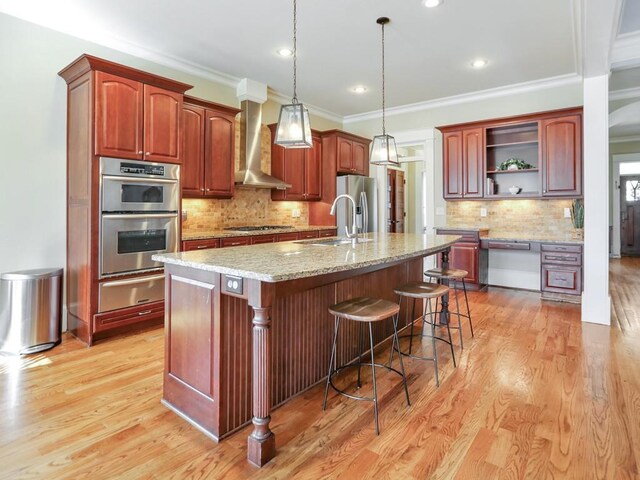 kitchen with wall chimney range hood, a kitchen breakfast bar, light stone counters, an island with sink, and decorative light fixtures