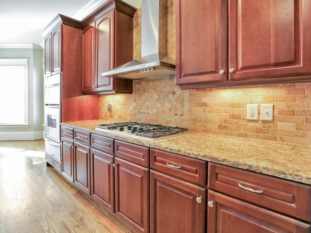 kitchen featuring tasteful backsplash, light stone counters, wall chimney exhaust hood, stainless steel gas cooktop, and crown molding