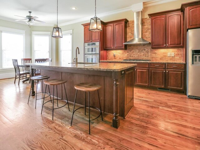 kitchen with a center island with sink, crown molding, wall chimney exhaust hood, appliances with stainless steel finishes, and stone countertops