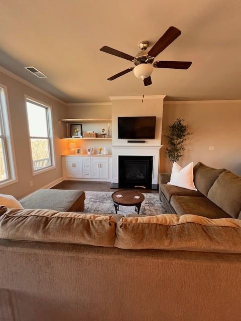 living area with ceiling fan, visible vents, baseboards, ornamental molding, and a glass covered fireplace