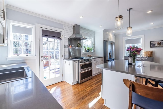 kitchen with light wood finished floors, crown molding, wall chimney range hood, stainless steel appliances, and white cabinetry