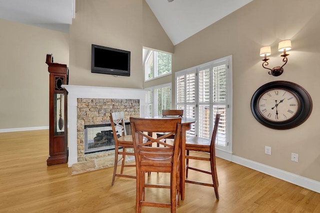 dining room featuring baseboards, light wood finished floors, a stone fireplace, and high vaulted ceiling