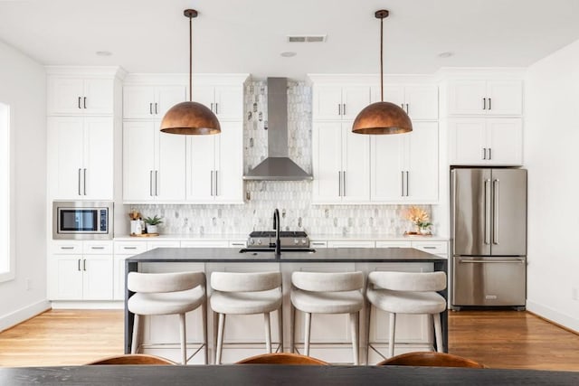 kitchen featuring backsplash, appliances with stainless steel finishes, a kitchen island with sink, and wall chimney range hood